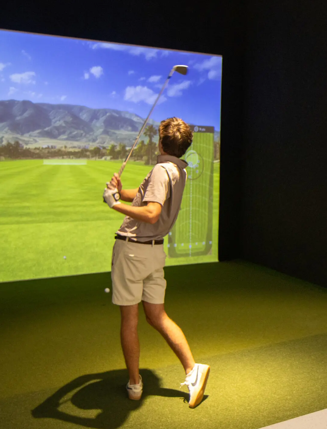 A student practices putting in a high-tech indoor golf simulator, with a large digital screen displaying a scenic golf course.